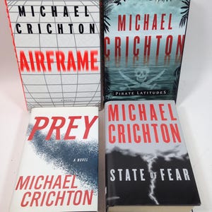 Può includere: Sono esposti quattro libri di Michael Crichton. I titoli includono "Airframe", "Pirate Latitudes", "Prey" e "State of Fear". Le copertine dei libri presentano vari design, tra cui una griglia, una scena tropicale, uno schizzo blu e un cielo tempestoso.