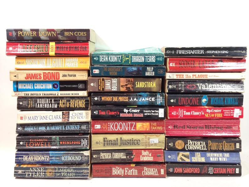 May include: A stack of hardcover books with various titles and authors. The spines display titles like "Power Down," "Firestarter," "James Bond," and "Sandstorm." The books are in various colors, including red, blue, and yellow.