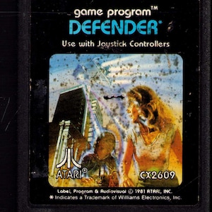 May include: Black Atari Defender video game cartridge. The label features the game title in teal, a futuristic cityscape, and a woman in a striped shirt. The Atari logo is visible. The cartridge is labeled CX2609.