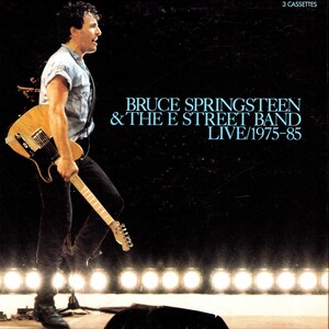 Puede incluir: Un set de casetes de música titulado Bruce Springsteen & The E Street Band Live/1975-85. La portada muestra a un artista con camisa clara y pantalones oscuros sosteniendo una guitarra eléctrica clara en un escenario con luces brillantes.