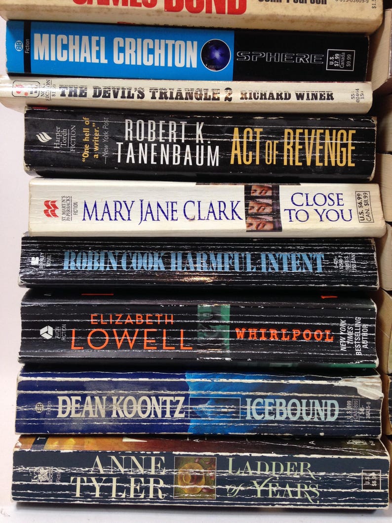 May include: A stack of paperback books with titles including "Sphere" by Michael Crichton, "Act of Revenge" by Robert K. Tanenbaum, and "Icebound" by Dean Koontz. The books have various colors and designs, with visible author names and book titles.
