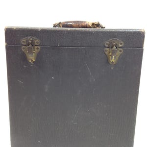 May include: A square, dark gray, textured carrying case with a worn brown leather handle on top. The case features two brass-colored metal latches on the front panel, showing signs of age and wear on the surface and hardware components.