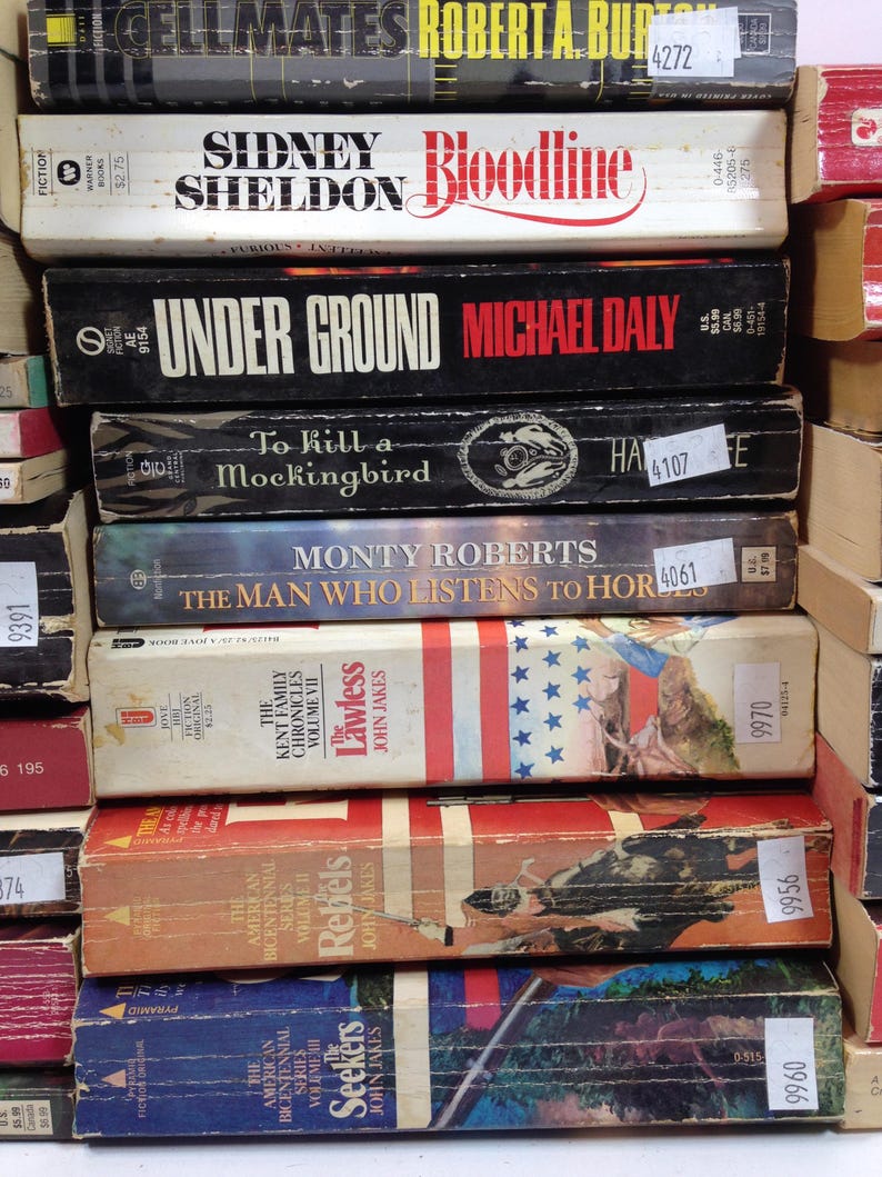 May include: A stack of vintage books with titles including "Cellmates," "Bloodline," and "To Kill a Mockingbird." The spines display various colors and fonts, with price tags and library stickers visible. The books are stacked together, suggesting a collection.