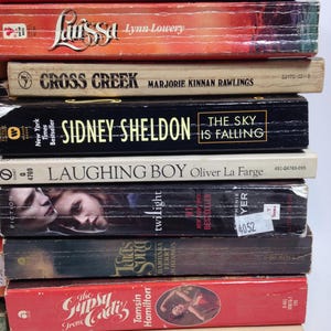 May include: A stack of vintage paperback books with titles including "Redeye," "Larissa," "Cross Creek," and "The Sky is Falling." The books have colorful covers and varying sizes, with visible author names.