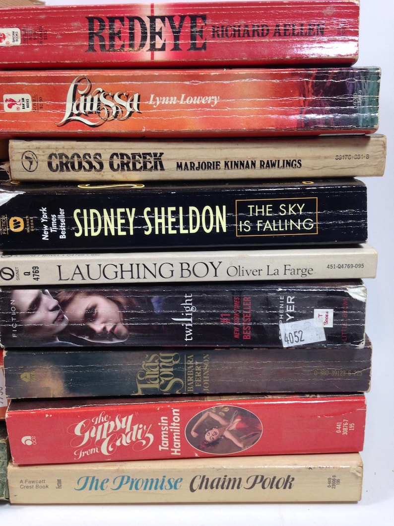 May include: A stack of vintage paperback books with titles including "Redeye," "Larissa," "Cross Creek," and "The Sky is Falling." The books have colorful covers and varying sizes, with visible author names.