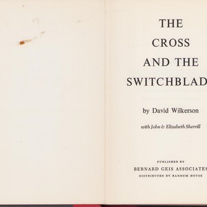 May include: Open book with the title "The Cross and the Switchblade" by David Wilkerson. The book's cover is off-white with black text. The publisher is Bernard Geis Associates.