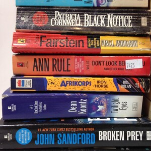 May include: A stack of paperback books with titles including "Dragon Tears," "Black Notice," and "Final Jeopardy." The books have colorful covers with author names and titles in bold text. The books are stacked vertically.