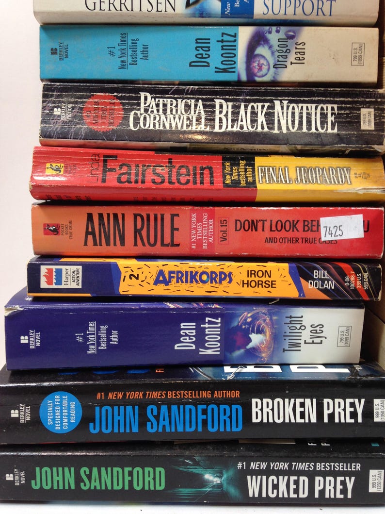May include: A stack of paperback books with titles including "Dragon Tears," "Black Notice," and "Final Jeopardy." The books have colorful covers with author names and titles in bold text. The books are stacked vertically.