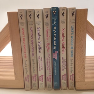 Puede incluir: Una colección de libros de bolsillo, incluyendo títulos como "Wyatt's Most Wanted Wife" y "Sky's Pride and Joy", sostenidos entre dos sujetalibros de madera. Los libros tienen lomos blancos con texto negro y un logotipo de silueta.