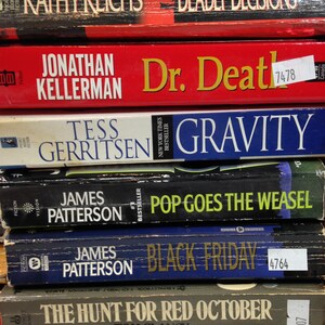 May include: A stack of hardcover books with titles including "Deadly Decisions," "Dr. Death," "Gravity," "Pop Goes the Weasel," "Black Friday," and "The Hunt for Red October." The books are in various colors, with visible author names.