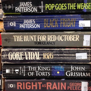 May include: A stack of hardcover books with various titles and authors visible. Titles include "Pop Goes the Weasel" by James Patterson, "Black Friday" by James Patterson, and "The Hunt for Red October" by Tom Clancy. The books are in various colors.