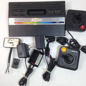 May include: A black Atari 2600 video game console with two black joysticks, power adapters, and a Gems-Tik joystick. The console has the Atari logo and the text "2600" on the front. The joysticks have red and yellow buttons.