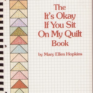 Könnte beinhalten: Ein spiralgebundenes Buch mit dem Titel The It's Okay If You Sit On My Quilt Book von Mary Ellen Hopkins, A Yours Truly Publication. Das Cover zeigt einen karierten Hintergrund und einen vertikalen Rand aus bunten, gemusterten Dreiecks-Quiltblöcken.