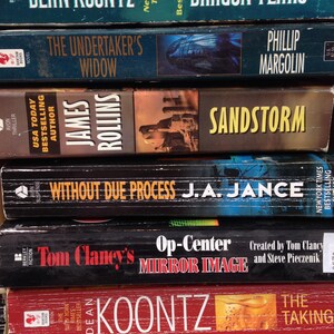 May include: A stack of paperback books with titles including "Dragon Tears," "The Undertaker's Widow," and "Sandstorm." The books have various colors and designs, with author names and book titles prominently displayed.