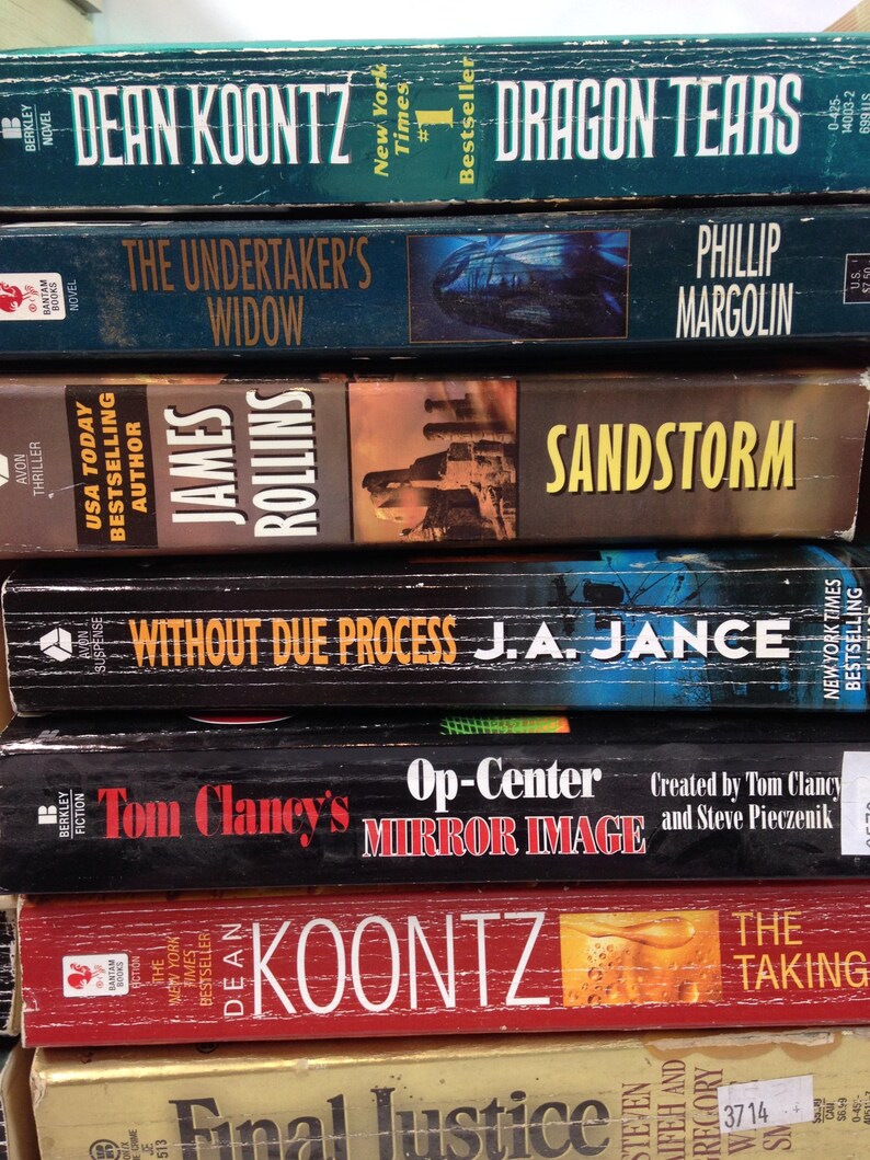 May include: A stack of paperback books with titles including "Dragon Tears," "The Undertaker's Widow," and "Sandstorm." The books have various colors and designs, with author names and book titles prominently displayed.