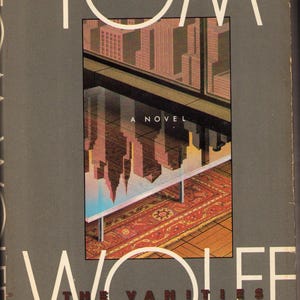 May include: A hardcover book titled The Bonfire of the Vanities by Tom Wolfe. The cover features a gray background with large white text and a central illustration of a city skyline reflected in a glass table surface above a patterned rug.