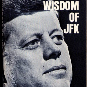 May include: Black and white book cover featuring a portrait of John F. Kennedy. The title "THE WISDOM OF JFK" is printed in large, white, bold letters across the top of the image. The image is a close-up of JFK's face.