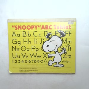 May include: A yellow "SNOOPY" ABC Tablet featuring a cartoon image of Snoopy, the beagle from the Peanuts comic strip, against a yellow background. The tablet has a lined surface with the alphabet and numbers printed on it. The text "SCHULZ" is printed in the bottom right corner.