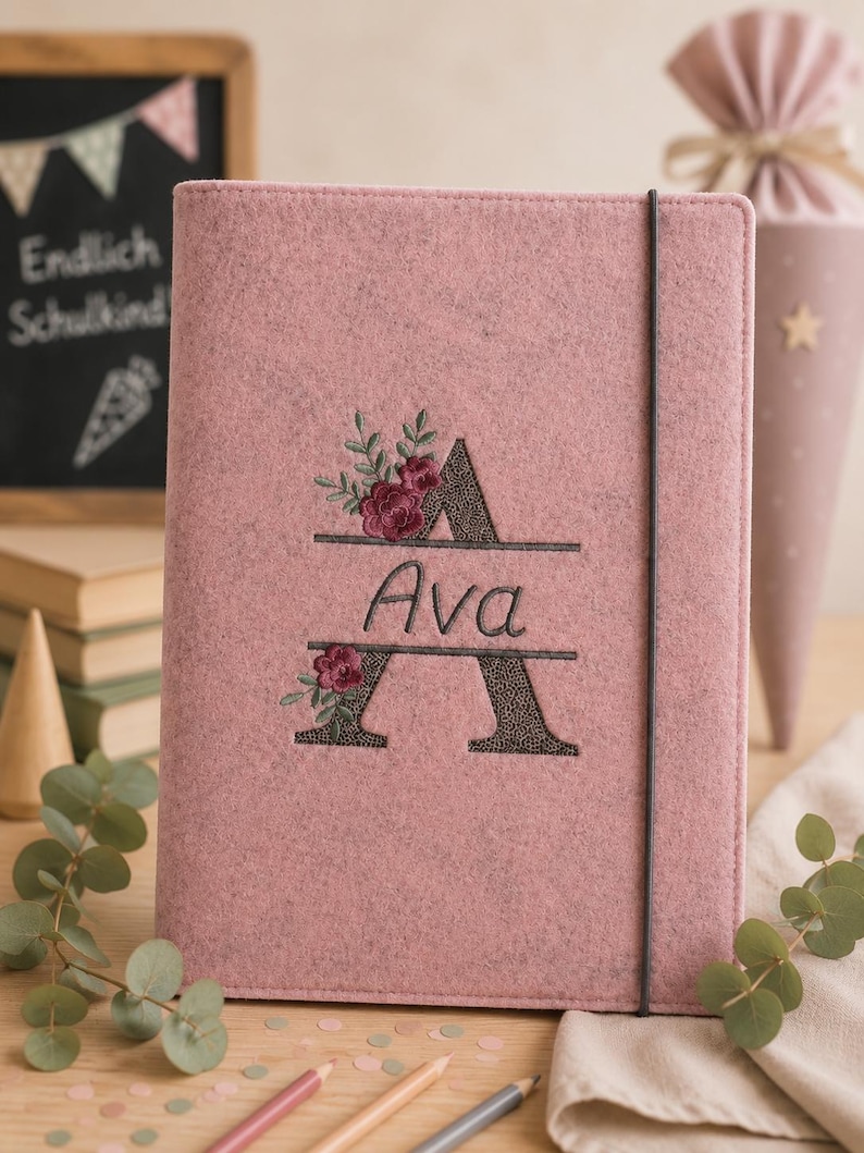 May include: A dusty pink felt notebook featuring a black elastic closure band. The cover is embroidered with a large letter A, split horizontally to display the name Ava in cursive script, accented with dark red floral and green leaf embroidery.