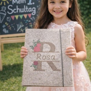 May include: A light beige felt folder featuring an embroidered large letter D and letter R with the name Rosa centered between them. The design includes pink and red floral accents with green leaves and a thin black elastic closure band.