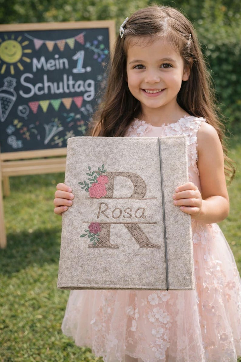 May include: A light beige felt folder featuring an embroidered large letter D and letter R with the name Rosa centered between them. The design includes pink and red floral accents with green leaves and a thin black elastic closure band.