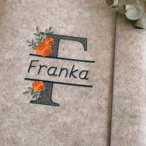 May include: A light beige felt folder featuring an embroidered dark green letter F with the name Franka stitched in the center. The design includes orange floral accents with green leaves. A thin gray elastic band secures the folder closed.