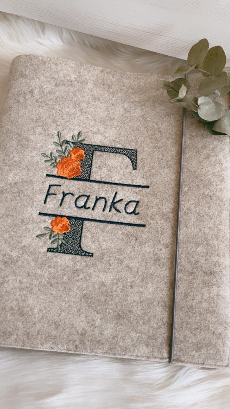 May include: A light beige felt folder featuring an embroidered dark green letter F with the name Franka stitched in the center. The design includes orange floral accents with green leaves. A thin gray elastic band secures the folder closed.