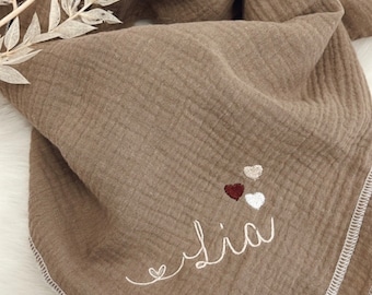May include: A brown muslin baby blanket with the name "Elia" embroidered in white thread. Three small embroidered hearts in brown, beige, and white are above the name.