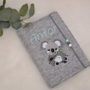 Könnte beinhalten: Ein grauer Filz-Notizbuchumschlag mit einem gestickten Koala, der einen Zweig hält, und dem Namen Anton in hellblauem Garn. Der Umschlag hat ein graues elastisches Verschlussband, das mit drei kleinen mintfarbenen, grauen und weißen Perlen verziert ist.