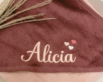 May include: A hooded baby towel in shades of mauve and light pink. The towel is embroidered with the name "Alicia" in a cursive font, accompanied by two small heart designs. The towel is folded, with a decorative sprig of dried grass.