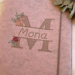 May include: A light pink felt folder featuring an embroidered design with the name Mona centered between two large letters M. The embroidery includes floral accents in shades of pink, orange, and green. A thin gray elastic band secures the folder.
