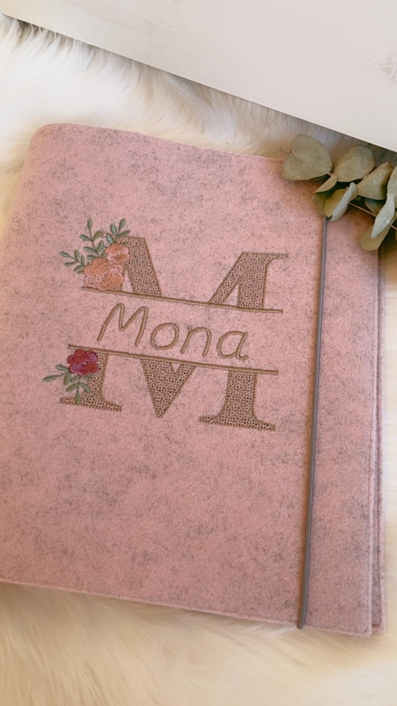 May include: A light pink felt folder featuring an embroidered design with the name Mona centered between two large letters M. The embroidery includes floral accents in shades of pink, orange, and green. A thin gray elastic band secures the folder.