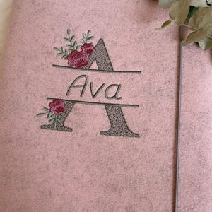 May include: A light pink felt folder featuring an embroidered design of a large letter A with a textured grey fill. The name Ava is stitched in black script across the center of the letter, accented by small embroidered pink floral sprigs.
