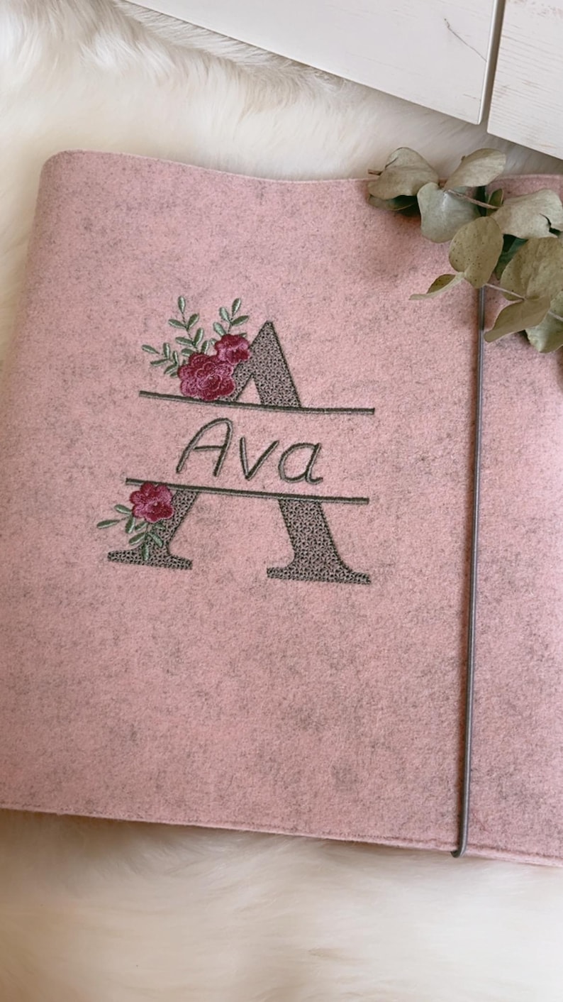 May include: A light pink felt folder featuring an embroidered design of a large letter A with a textured grey fill. The name Ava is stitched in black script across the center of the letter, accented by small embroidered pink floral sprigs.