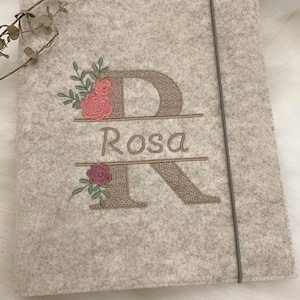 May include: A light gray felt folder featuring an embroidered design of a large letter R with the name Rosa stitched in the center. The embroidery includes pink and dark red floral accents with green leaves and a thin gray elastic closure band.
