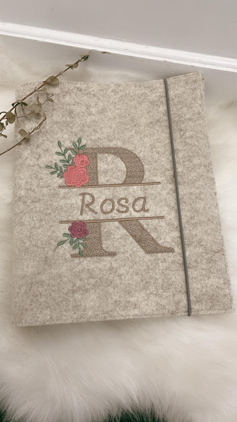 May include: A light gray felt folder featuring an embroidered design of a large letter R with the name Rosa stitched in the center. The embroidery includes pink and dark red floral accents with green leaves and a thin gray elastic closure band.