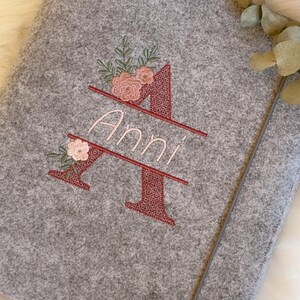 May include: A grey felt notebook cover featuring an embroidered large letter A in a dark red textured stitch. The name Anni is embroidered in light pink script across the center of the letter, accented with small pink flowers and green leaves.