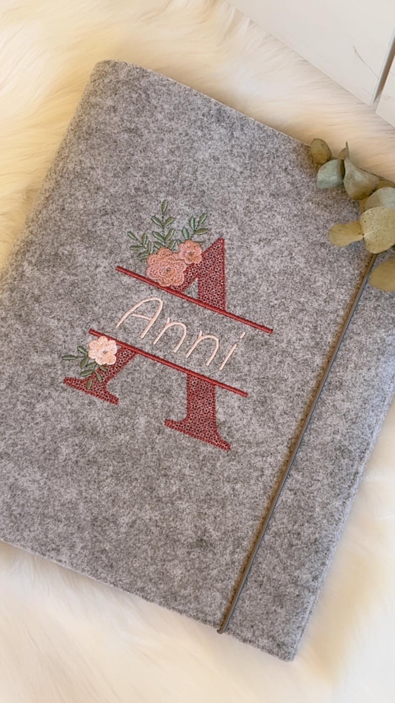 May include: A grey felt notebook cover featuring an embroidered large letter A in a dark red textured stitch. The name Anni is embroidered in light pink script across the center of the letter, accented with small pink flowers and green leaves.