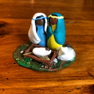 May include: A clay nativity scene with a white baby Jesus in a manger, a blue and white robed woman, and a yellow and blue robed man. The scene is set on a green and brown clay base with white cotton and small white eggs.
