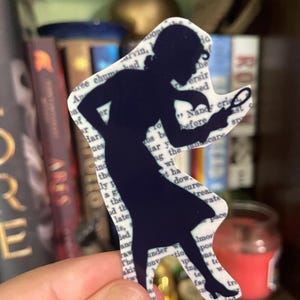 May include: A navy blue silhouette of a woman holding a magnifying glass, cut out in the shape of the figure. The cutout is layered over a background of book pages. The figure is likely a detective.