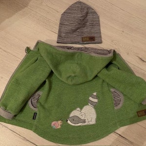 Könnte beinhalten: Grüner Kapuzenpullover mit einem Waschbär- und Schnecken-Applikationen. Der Pullover hat einen gestreiften grauen und weißen Besatz um die Kapuze und die Taschen. Der Text "Little King" befindet sich auf der rechten Seite des Pullovers.