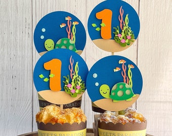May include: Four cupcake toppers with a blue ocean background, a green turtle, orange number one, and coral reef decorations. The toppers are on top of cupcakes with brown wrappers that say "Sweets Paradise".