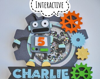 May include: A robot-themed cake topper with the name "Charlie" in blue letters on a black banner. The topper features a gray robot with a number 5 on its chest, surrounded by colorful gears and the word "INTERACTIVE".