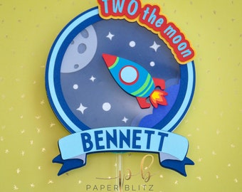 May include: A blue and white cake topper with a rocket ship flying through a starry night sky. The topper says "TWO the moon" and "BENNETT" in a blue banner.