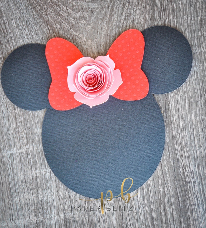 以下が含まれることがあります： 黒い紙で作ったミッキーマウスの耳の切り抜き。赤いリボンとピンクの紙の花が上に飾られています。画像の下には「PAPER BLITZ」という文字があります。