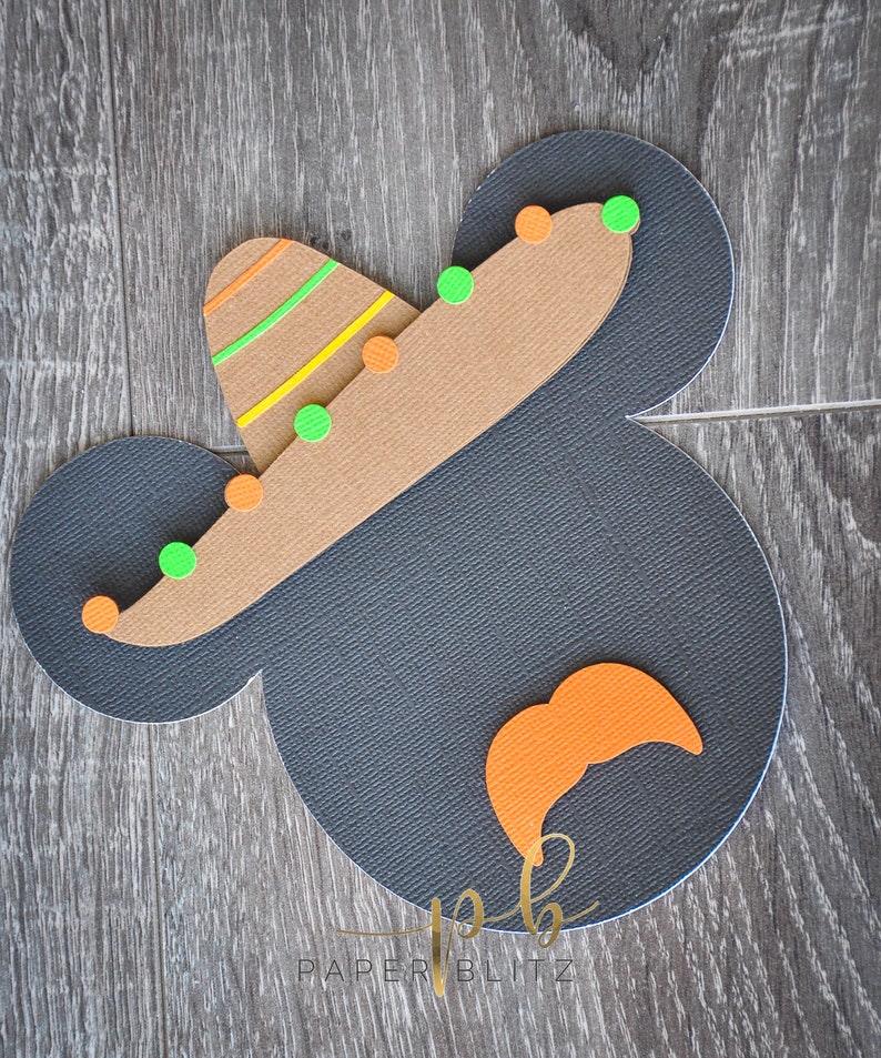 以下が含まれることがあります： 黒と茶色の紙でできたミッキーマウスの頭のクラフトで、緑とオレンジのアクセントがついた茶色のソンブレロをかぶっています。ソンブレロには黄色と緑のストライプがあります。クラフトは木目調の背景の上にあります。画像の下部には金色の文字で「Paper Blitz」と書かれています。