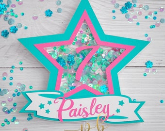 May include: A teal and pink star-shaped cake topper with a clear window filled with iridescent sequins. The number 7 is visible in the window. A white banner with the name "Paisley" and two stars is attached to the bottom of the star. The text "Paper Blitz" is printed below the banner.