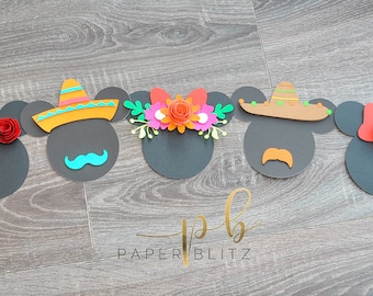 May include: Seven paper cutouts of Mickey Mouse heads with different Mexican-themed decorations. The decorations include a sombrero, a flower crown, a red bow, and a mustache. The cutouts are on a woodgrain background.