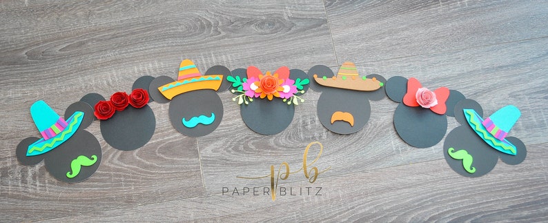 以下が含まれることがあります： メキシコをテーマにした装飾が施されたミッキーマウスの頭部の紙切りが7つあります。装飾には、ソンブレロ、花冠、赤いリボン、口ひげがあります。切り抜きは木目調の背景に置かれています。