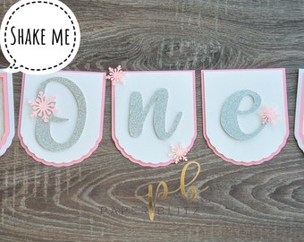 May include: A banner for a first birthday celebration. The banner features the word "one" spelled out in silver glitter letters, with a polar bear on each end. The banner is white with pink accents and snowflake details.